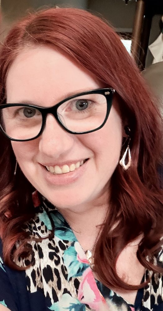 Terri Ellis, smiling, with red hair and black cat-eye glasses